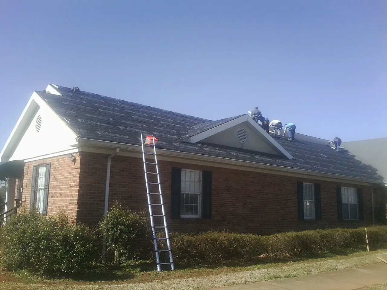 Crew tearing shingles off a brick ranch roof with a ladder set — Emergency Roof Repair work by our crew in Ware Shoals
