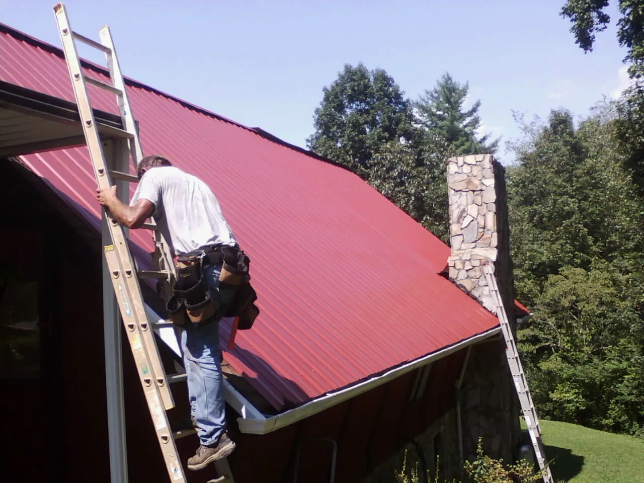 Roofer on a ladder beside a red metal roof and stone chimney — Soffit & Fascia Repair by Roofing Brothers in Ware Shoals
